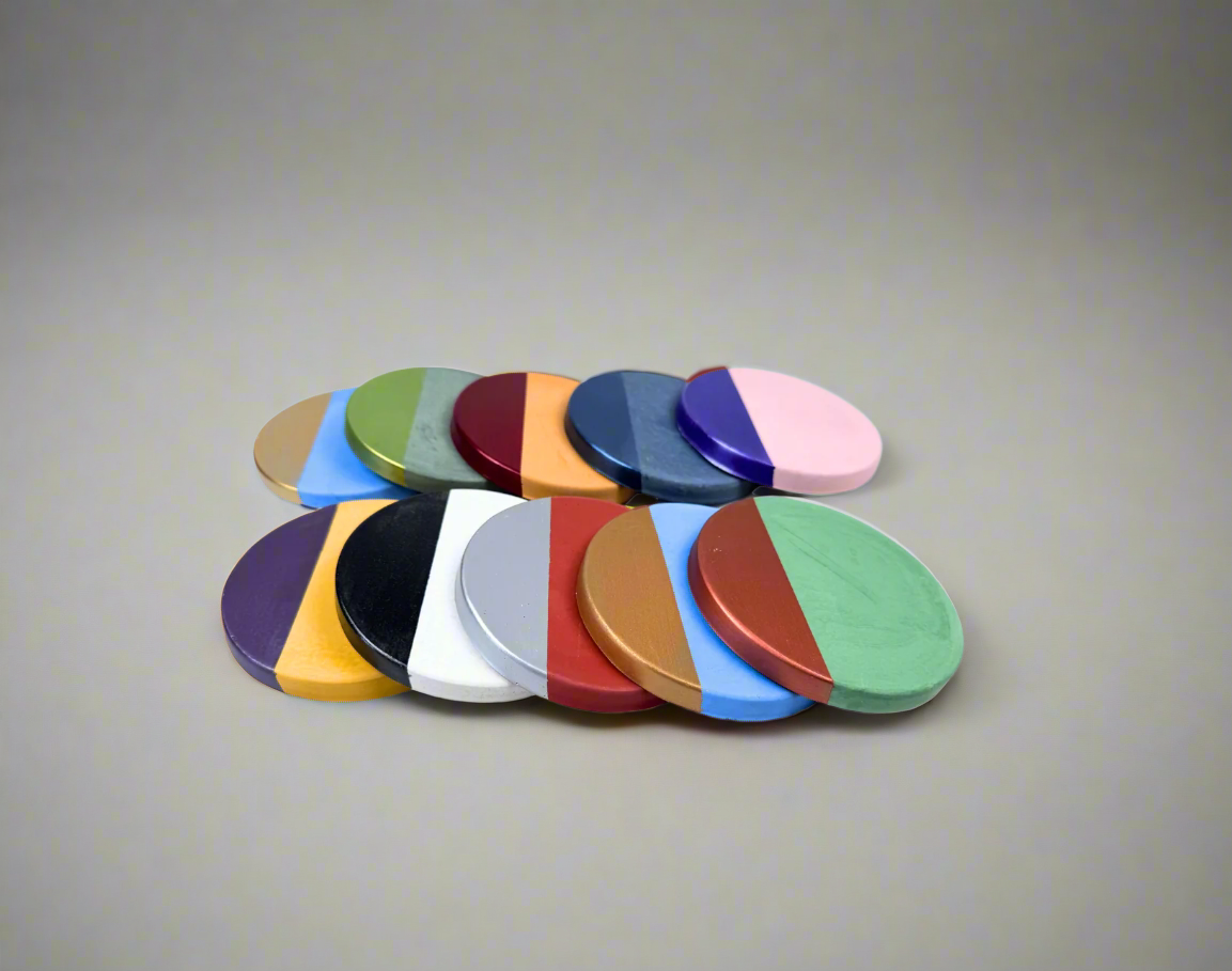 A close-up overhead shot showcases a variety of round concrete coasters, each with a cement base color and a contrasting vibrant or metallic accent color hand-painted in a semi-circular design, creating a personalized and colorful effect against a white background.
