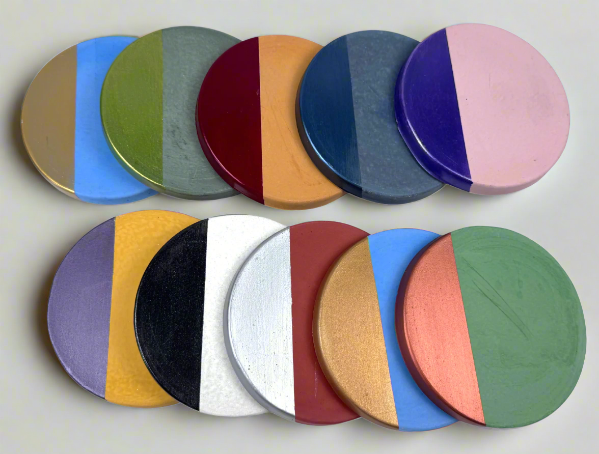 A collection of round concrete coasters is arranged in two rows on a white background. Each coaster features a distinct two-tone design with a base color and a contrasting semi-circular accent hand-painted in various vibrant and metallic shades, showcasing the wide range of personalization available.