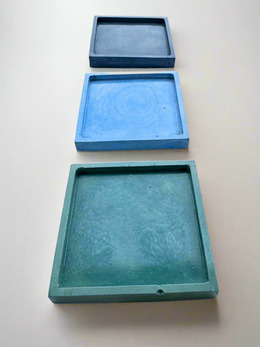 Three glossy square concrete coaster trays in Dark blue, Light Blue, and Turquoise are arranged in a vertical line on a white surface, showcasing the subtle lip designed to catch spills.