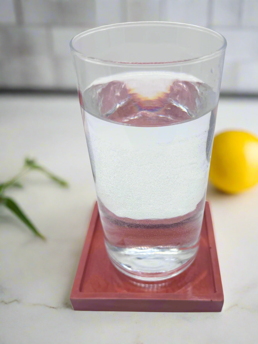 A clear glass of water sits on a glossy Barn Red square concrete coaster tray on a white surface, with a blurred black and red bowl visible in the background.