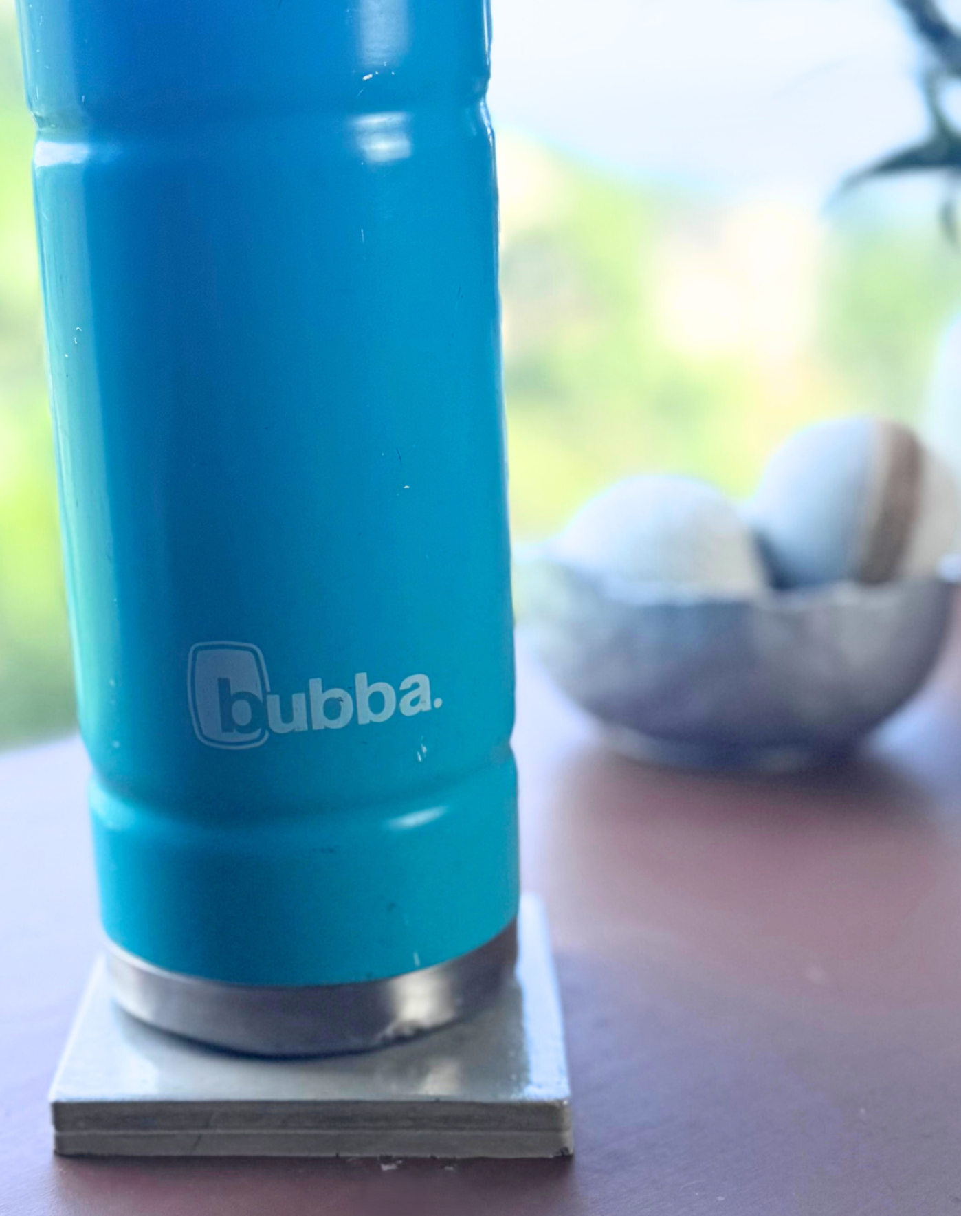 A bright green "Bubba" water bottle rests on a glossy Cement Grey square concrete coaster on a wooden surface, with a blurred background showing a metallic bowl and white decorative balls.