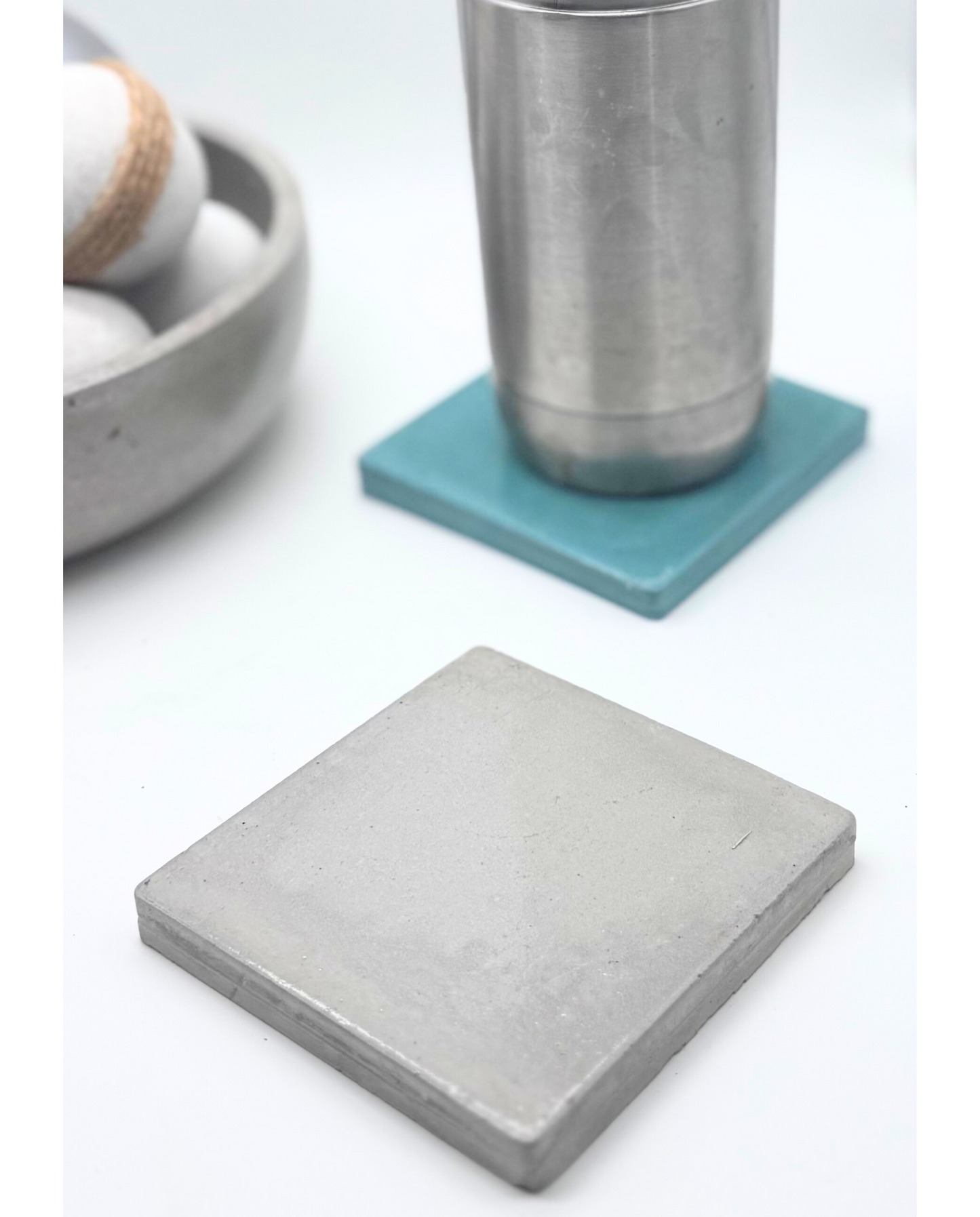 A glossy Cement Grey square concrete coaster is in the foreground, with a stainless steel cup on a glossy Turquoise coaster and a concrete bowl with white and twine-wrapped spheres blurred in the background.