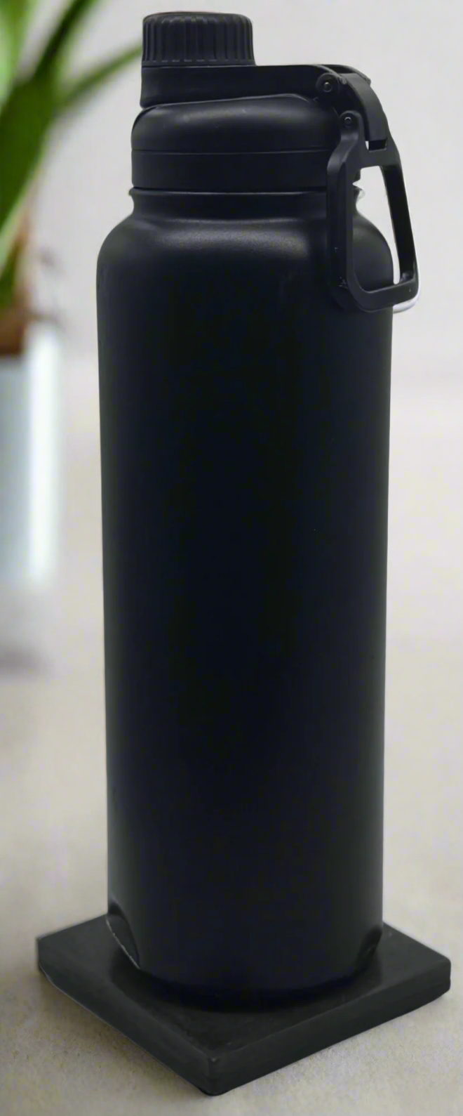 A tall black water bottle with a black lid and handle rests on a glossy Charcoal square concrete coaster against a white background.