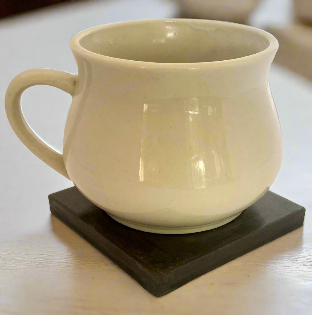 An Ivory White ceramic mug with a handle sits on a glossy Charcoal square concrete coaster against a white background.