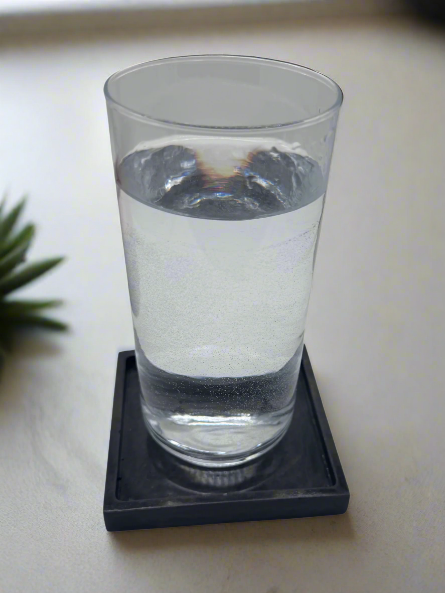 A clear glass filled with water rests on a glossy Charcoal square concrete coaster tray, highlighting the subtle lip designed to contain condensation, all against a white background.