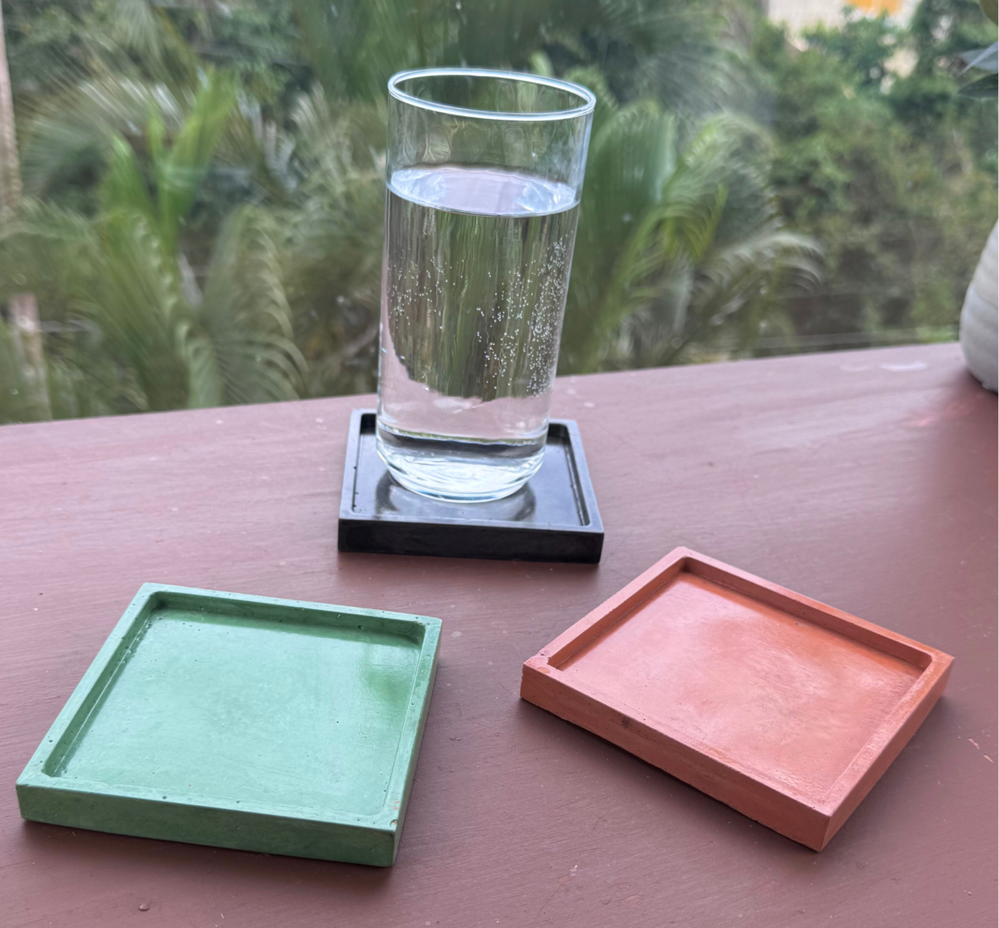 Three glossy square concrete coaster trays in Charcoal, Green, and Coral are arranged on a brown surface with a blurred outdoor background; a clear glass of water sits on the Charcoal tray, illustrating its use as a spill-catching drink holder.
