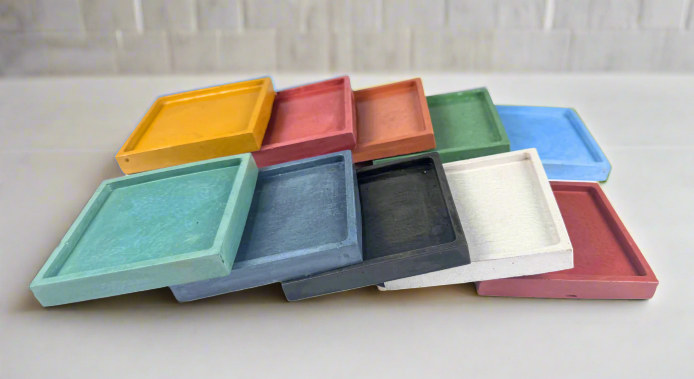en glossy square concrete coaster trays in various colors including Mustard Yellow, Barn Red, Coral, Green, Light Blue, Turquoise, Dark Blue, Charcoal, Ivory White, and Mauve are stacked and slightly overlapping on a white background, showcasing the range of available shades for these spill-catching drink holders.