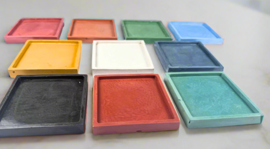 Twelve glossy square concrete coaster trays in various colors including Barn Red, Coral, Green, Light Blue, Honey Yellow, Ivory White, Charcoal, Blush Pink, Dark Green, Dark Blue, Mustard Yellow, and Turquoise are scattered and slightly overlapping on a white background, showcasing the range of available shades for these spill-catching drink holders.