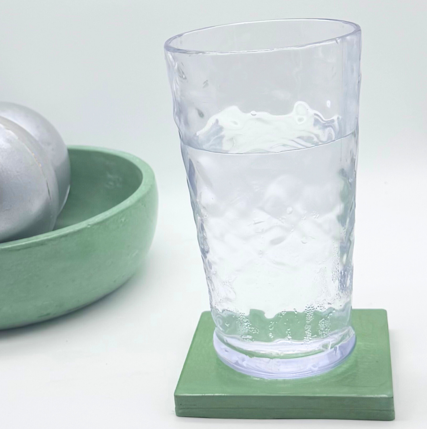 A clear glass of water rests on a glossy Green square concrete coaster, with a pale green concrete bowl containing two metallic spheres partially visible in the blurred background.