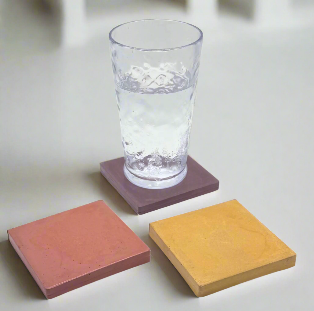 A clear glass of water sits atop a glossy Mauve square concrete coaster, flanked by a Mustard Yellow and a Coral coaster on a white background.