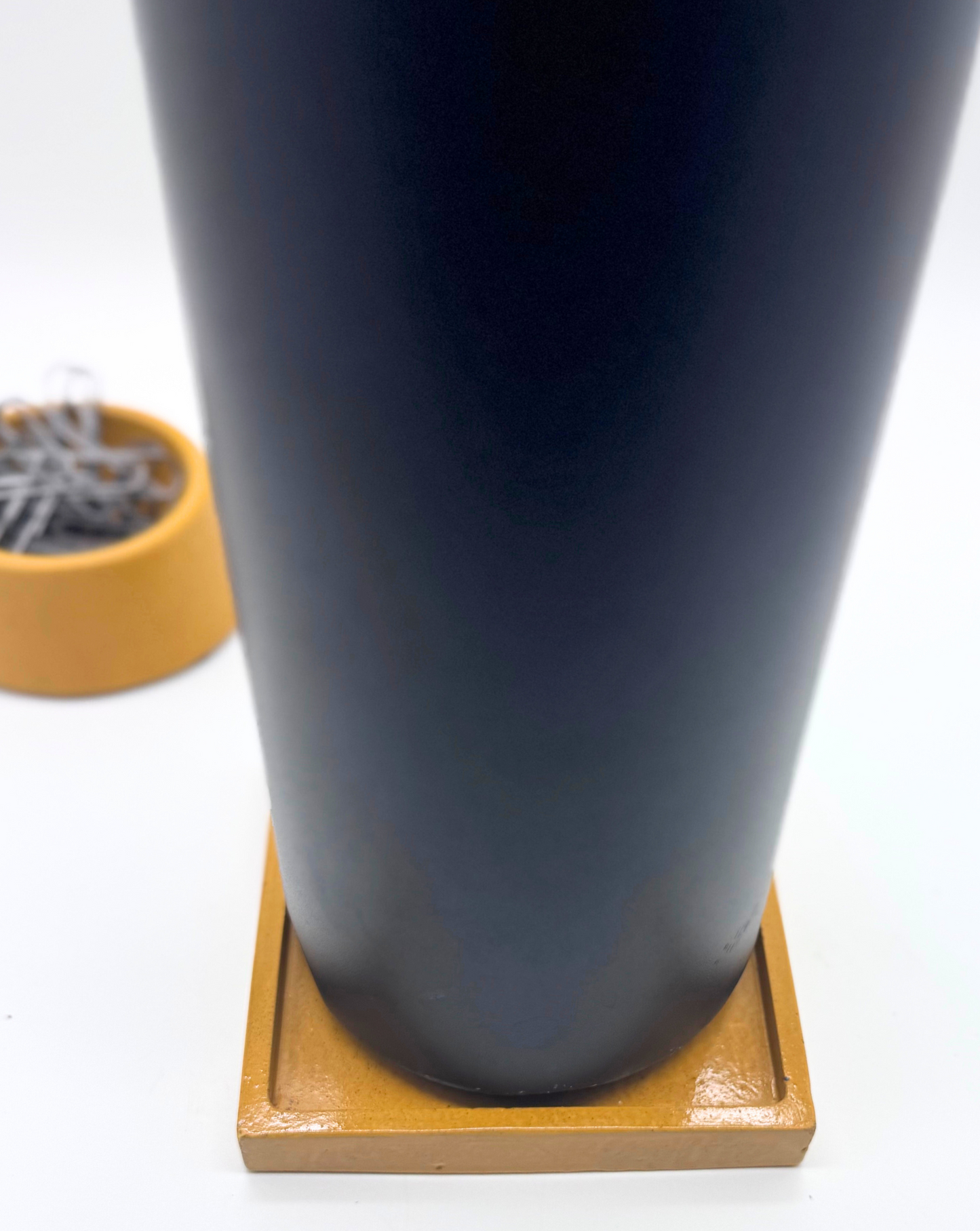 A tall black tumbler rests on a glossy Mustard Yellow square concrete coaster tray on a white surface, with a blurred orange magnetic paperclip holder containing silver paperclips visible in the background.