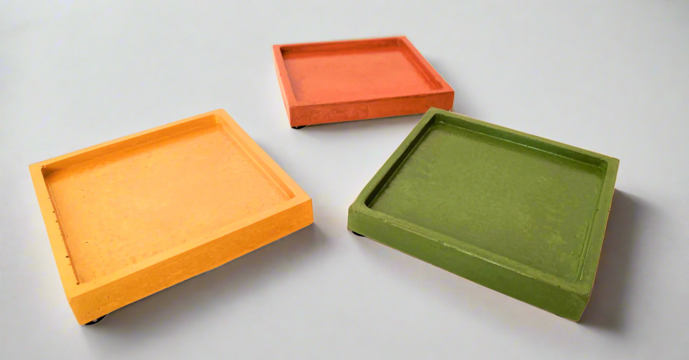 Three glossy square concrete coaster trays with black felt pads on the bottom, in Mustard Yellow, Coral, and Green, are angled on a white background.