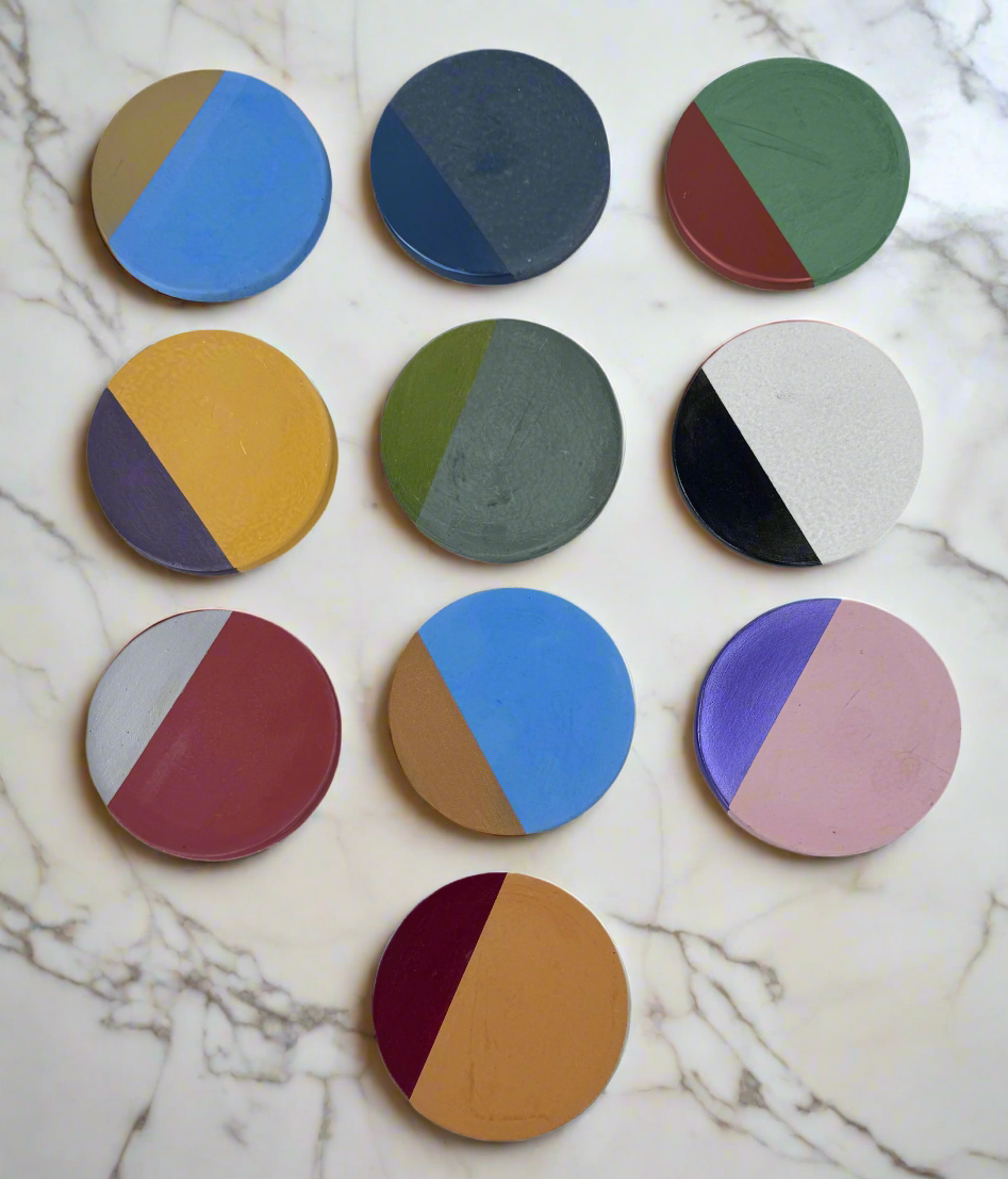 Ten round concrete coasters are organized in a grid on a white background. Each coaster has a cement base color and a contrasting hand-painted semi-circular accent in various metallic and vibrant colors, illustrating the customizable design of these coasters.