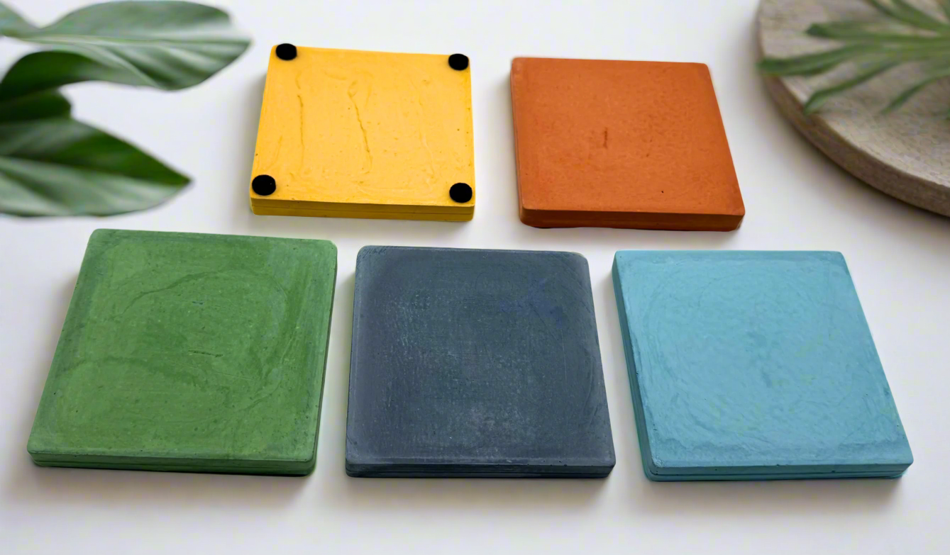 The image displays five coasters set against a clean white background. The coasters are presented in a variety of colors: mustard yellow, squash orange, green, dark blue, and light blue. The mustard yellow coaster is distinctively placed upside down, showing its underside which features four felt pads.