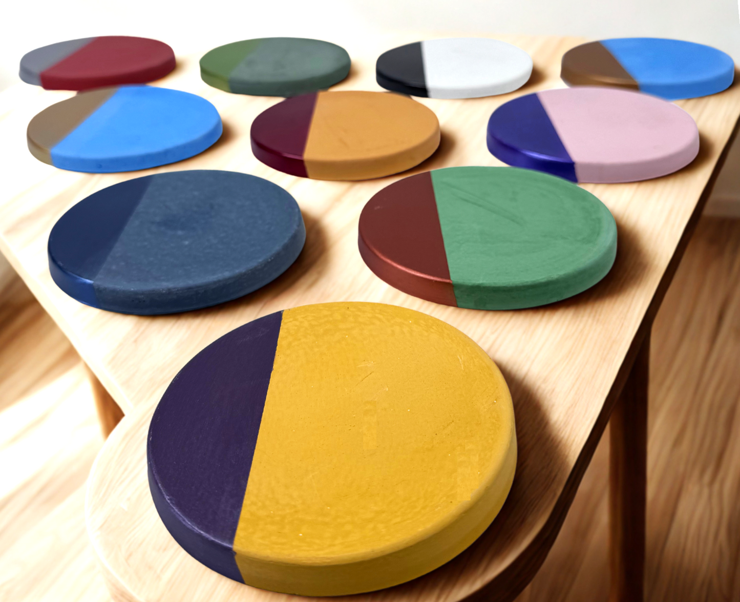 Ten round concrete coasters, each with a unique hand-painted accent color on one half, are displayed on a light-colored wooden surface, highlighting the variety of customization options for these personalized drink coasters.