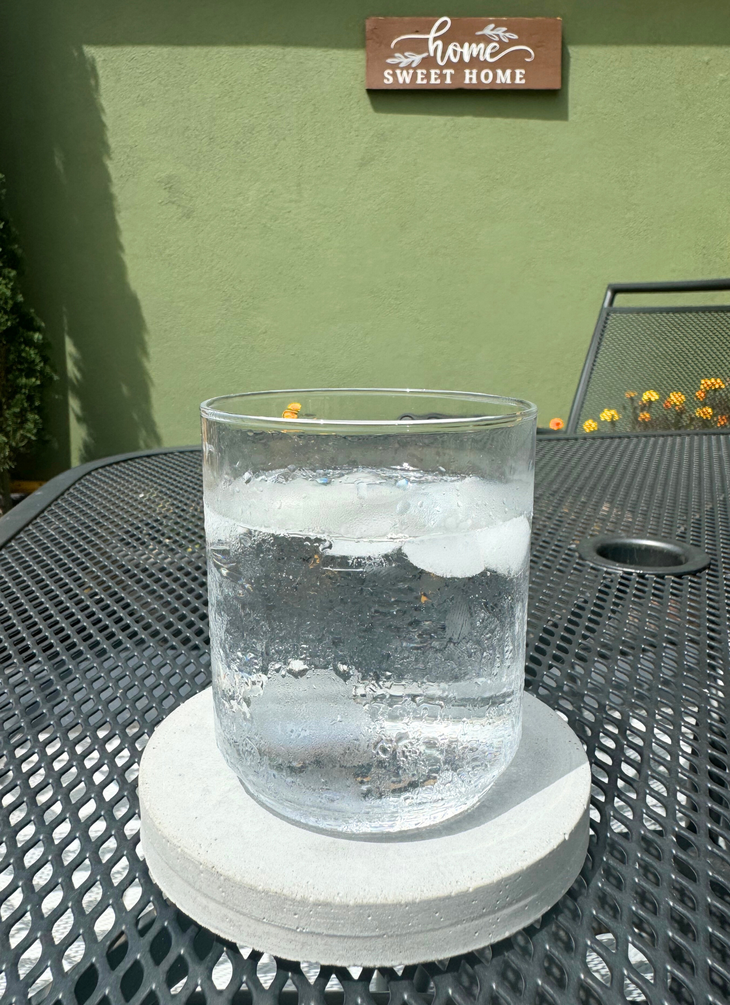 Round cement gray concrete coaster holding a glass on an outdoor patio table. "Home Sweet Home" sign visible in background.