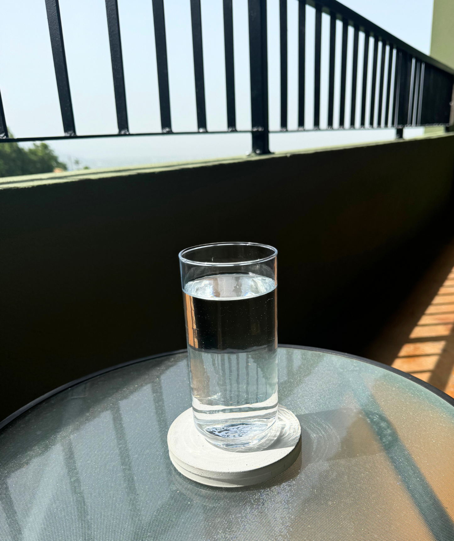 Round cement gray concrete coaster holds a glass of water on a glass patio table. Sky, railing, and shadows visible in background.