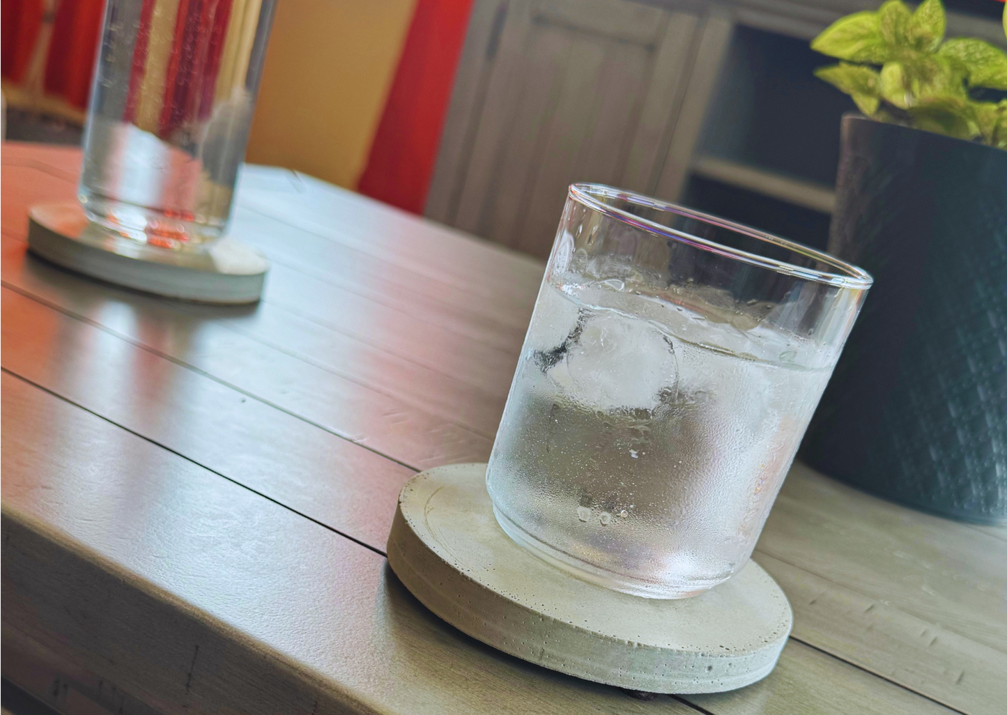 Round cement gray concrete coaster holding an iced glass on a coffee table. Blurred background features a plant and another coaster.