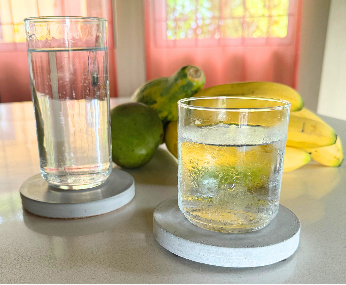 Two round cement gray concrete coasters hold tall & short glasses on white kitchen counter. Blurred fruits & window view behind