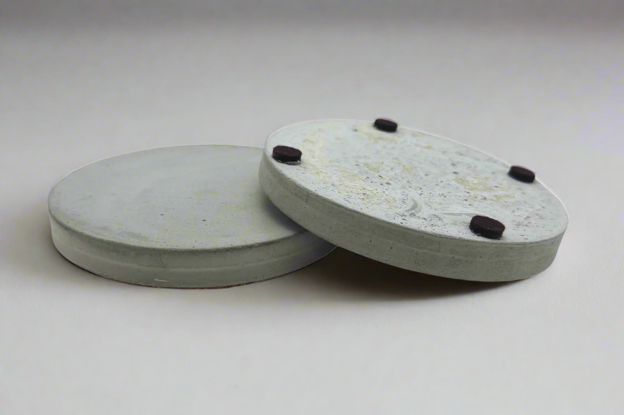 Two round cement gray concrete coasters. One lays flat, the second rests tilted showing four protective felt pads on its base.