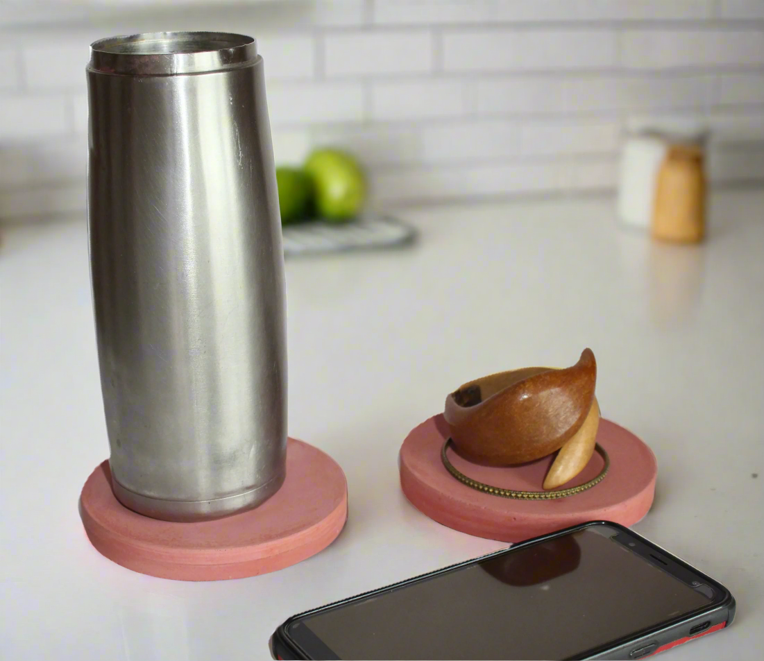 Two round handmade concrete drink coasters are on a white surface: a stainless steel tumbler rests on a Blush Pink coaster, while a small wooden bracelet holder with a bracelet sits on a Barn Red coaster; a black smartphone is partially visible in the foreground.