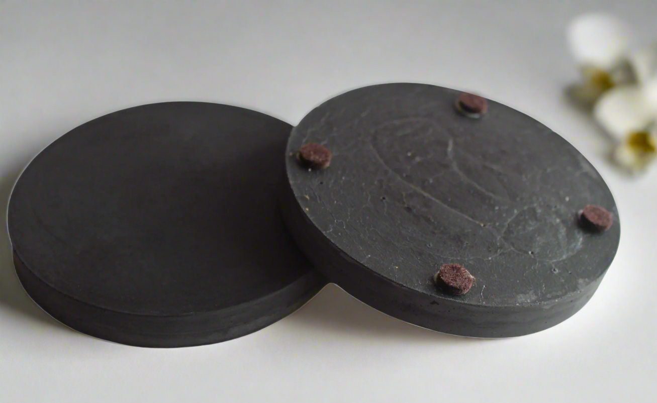 Two round handmade concrete drink coasters in Charcoal are shown against a white background, one lying flat and the other slightly tilted to reveal four small brown felt feet on its underside, highlighting the protective base option.