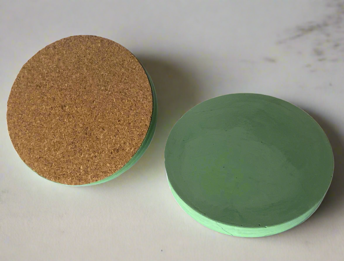 Two round handmade concrete drink coasters in Green are displayed side-by-side on a white background. The coaster on the left is flipped to show its cork backing, highlighting the protective base option, while the coaster on the right is face-up.