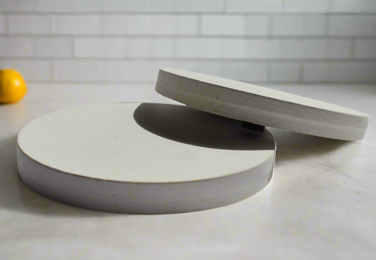 Two round handmade concrete drink coasters in Ivory White are slightly stacked against a white background, one lying flat and the other leaning on top, revealing their circular shape and substantial thickness.