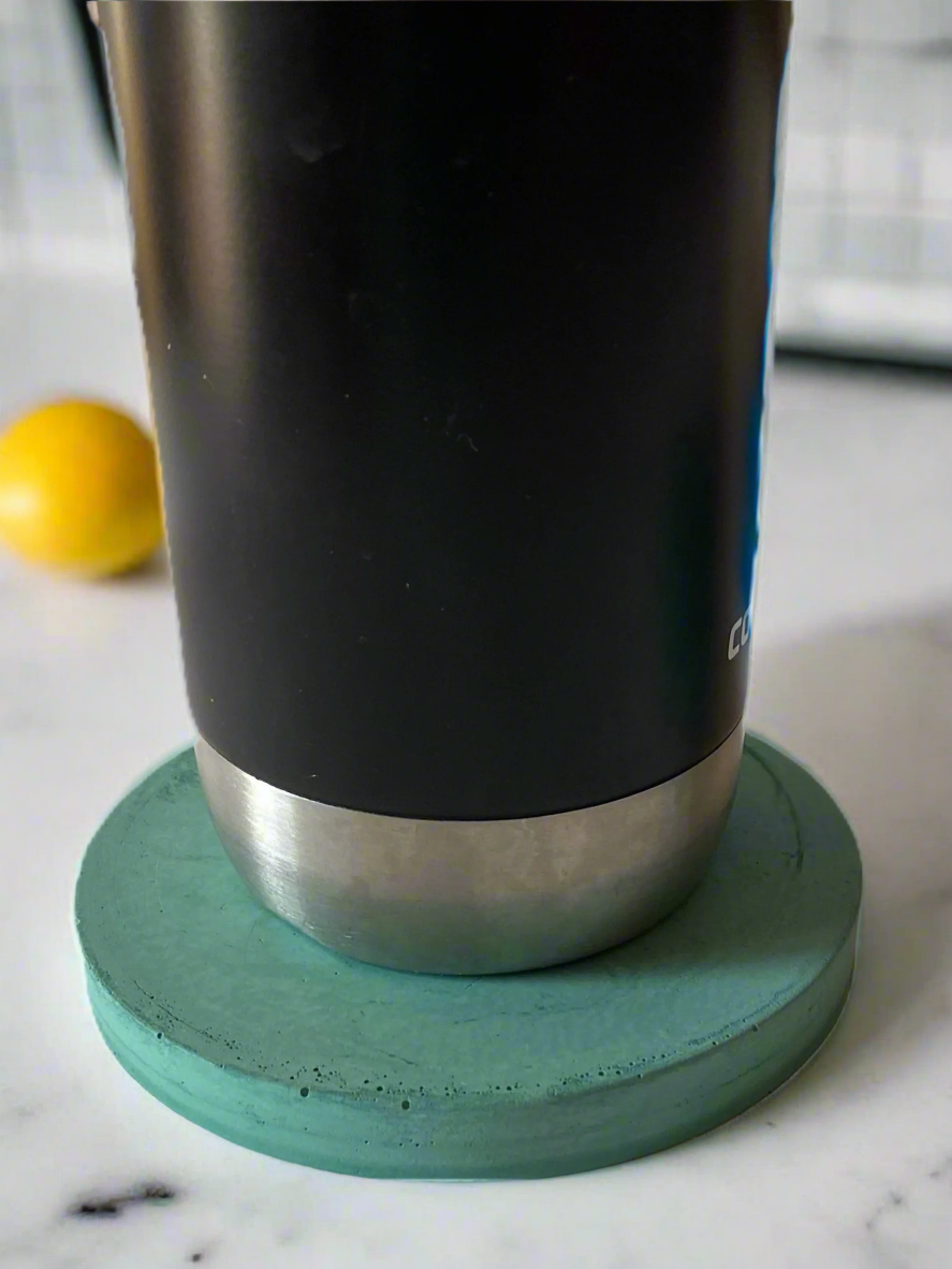 A black tumbler with a silver base rests on a round handmade concrete drink coaster in Turquoise against a white background, showcasing the coaster's texture and circular form.