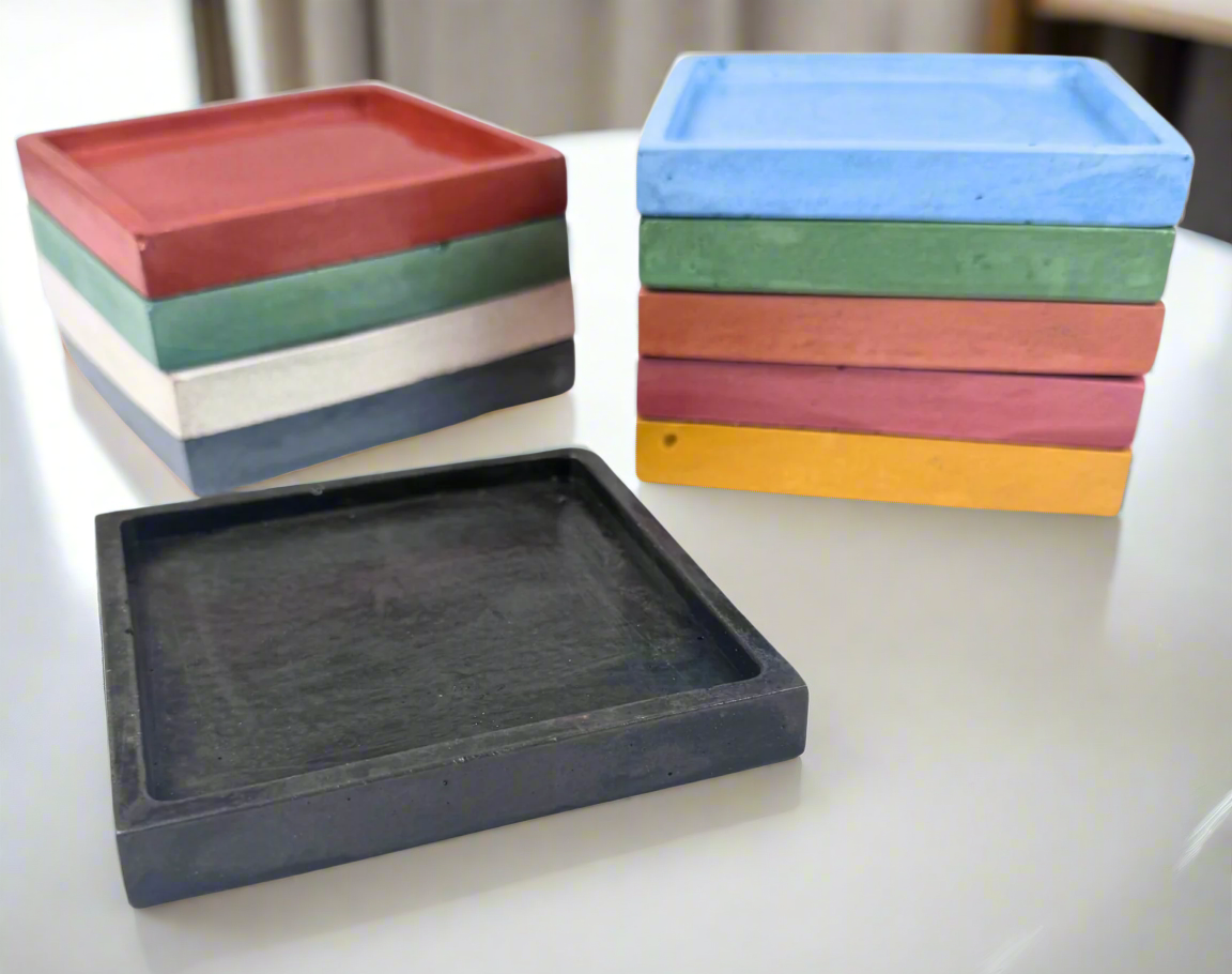Two stacks of glossy square concrete coaster trays, one with five trays in Barn Red, Coral, Green, Ivory White, and Dark Blue, and the other with five trays in Light Blue, Green, Coral, Barn Red, and Honey Yellow, sit on a white background, with a single Charcoal tray in the foreground, showcasing the variety of colors available for these spill-catching drink holders.